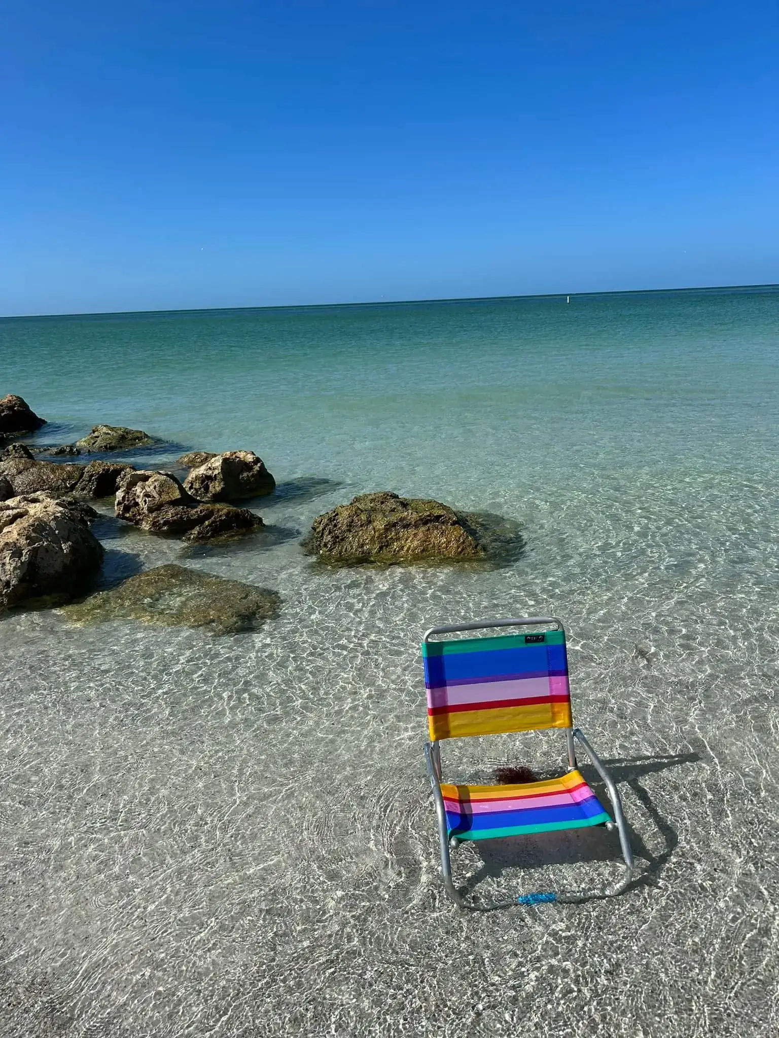 Colorful beach chair on clear sandy shore with rocks and calm sea.