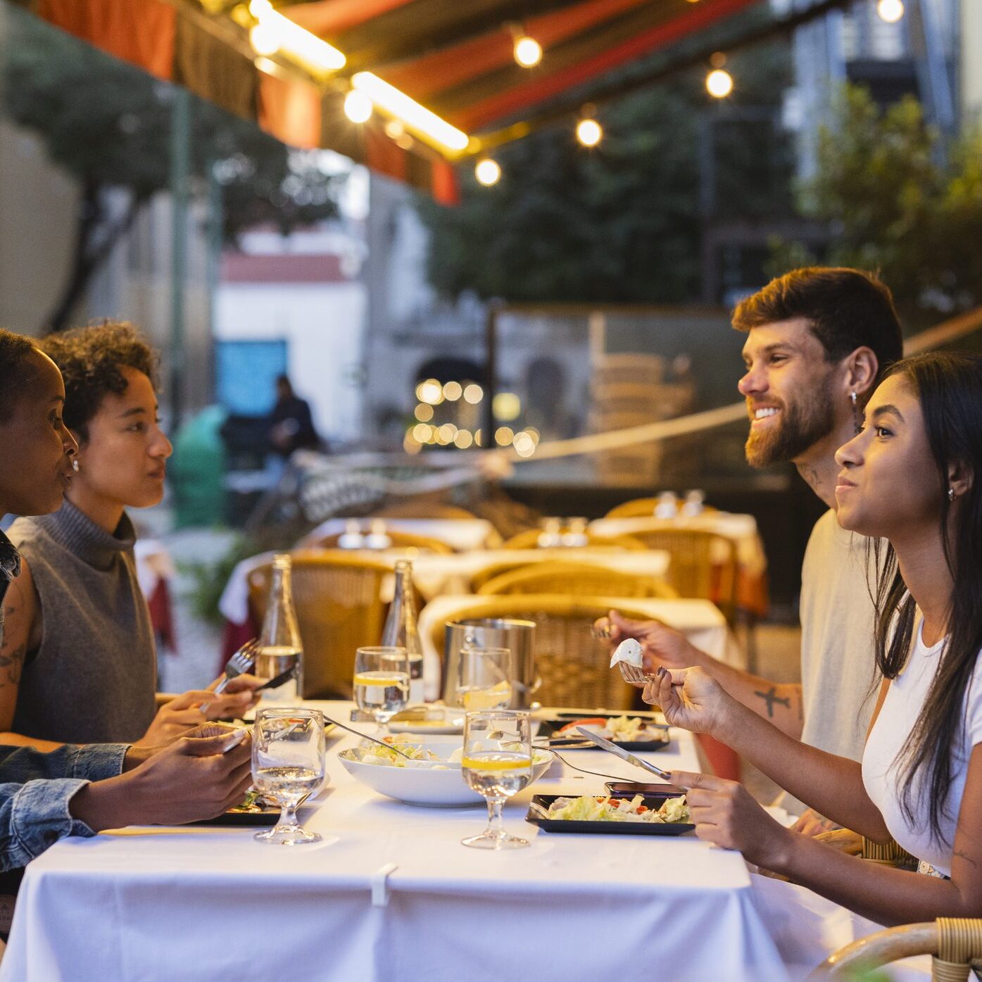 Four friends enjoying an outdoor dinner at a restaurant under warm lighting.