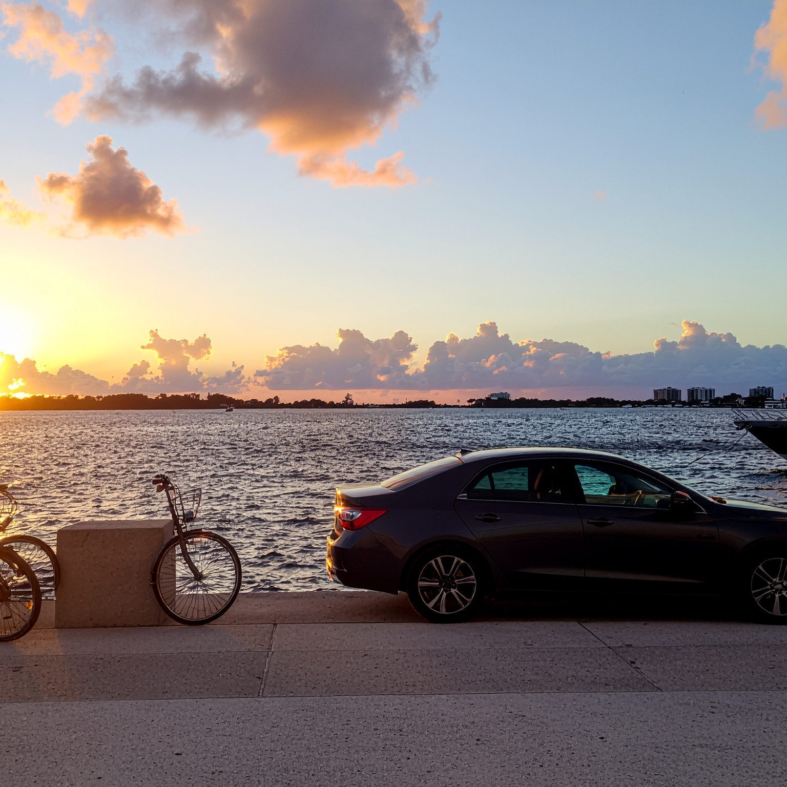A bicycle and parked car by the waterfront at sunset.